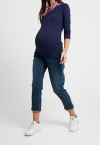 Laivastonsininen pitkähiha -paita, jossa on rypistetyt sivut, yhdistettynä käännettävillä hihoilla varustettuihin farkkuihin ja valkoisiin tennareihin. Värikäs kuhmuinen huivi lisää yksityiskohtia.