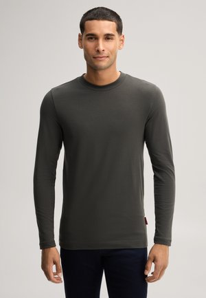 Man met kort haar die een donkergroen shirt met lange mouwen en ronde hals draagt, staand tegen een effen lichte achtergrond.