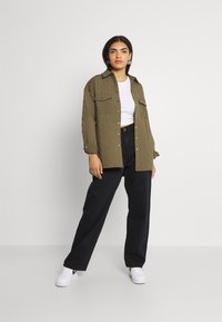 Camisa acolchoada verde-oliva com dois bolsos no peito, usada sobre uma blusa branca, combinada com calças largas pretas e ténis brancos.