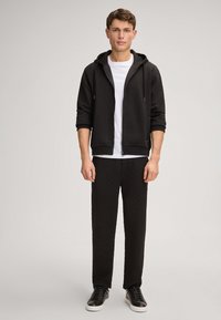 Felpa nera con zip e finitura texturizzata, abbinata a pantaloni coordinati. Presenta una t-shirt bianca sotto e sneakers nere.