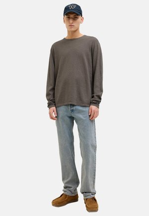 Giovane uomo in piedi, indossa una maglia a maniche lunghe grigio scuro, jeans azzurri, scarpe di camoscio marrone e un cappellino blu navy con un logo bianco a forma di "W".