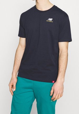 Dunkelblaues Baumwoll-T-Shirt mit kurzen Ärmeln, versehen mit einem weißen Logo und goldenem Schriftzug. Kombiniert mit türkisfarbenen Jogginghosen, die ein rotes Etikett zeigen.