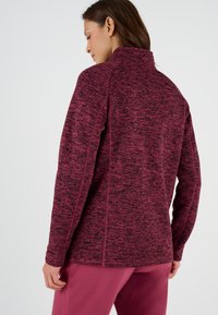 Chaqueta de forro polar burdeos con cuello alto, tejido texturizado y costuras laterales. La espalda presenta una silueta limpia y moderna sin cierres.