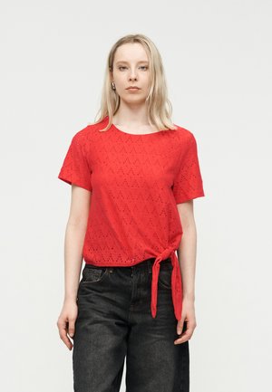 Kvinde iført en rød kortærmet top med sidelukning og sorte højtaljede jeans, stående mod en ensartet lys baggrund.