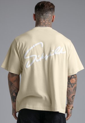 Man met tatoeages op nek en arm draagt een beige oversized T-shirt met groot wit scriptlogo op de rug, staand tegen een grijze achtergrond.