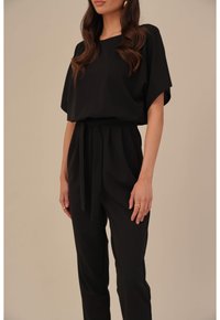 Femme portant un haut noir à manches courtes amples et un pantalon noir taille haute avec une ceinture en tissu, se tenant devant un fond beige.