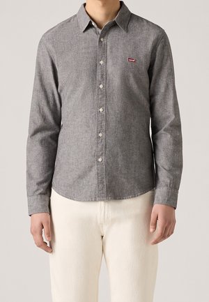 Homme portant une chemise grise à manches longues avec un petit logo rouge sur la poitrine, associée à un pantalon écru, debout devant un fond uni.