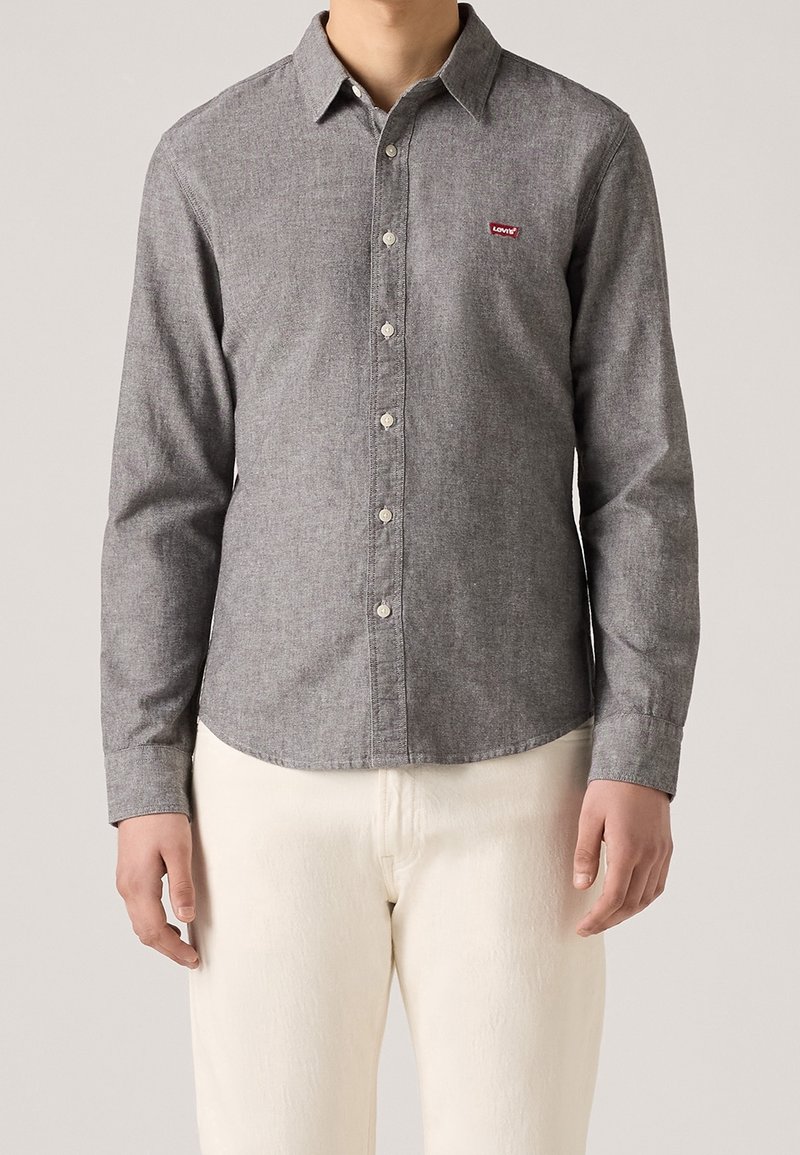 Homme portant une chemise grise à manches longues avec un petit logo rouge sur la poitrine, associée à un pantalon écru, debout devant un fond uni.