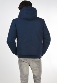 Mørkeblå polstret jakke med hætte, med en glat tekstur og elastiske manchetter, båret med lysegrå denim jeans.