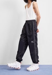 Schwarze Nike-Trainingshose mit weißen Seitenstreifen und Logo, kombiniert mit weißen High-Top-Sneakern mit roten Akzenten. Glatter, leichter Stoff.