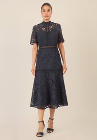 Abito in pizzo blu navy con motivi floreali, maniche corte, scollatura alta e gonna svasata. Decorato con rifiniture e intagli.