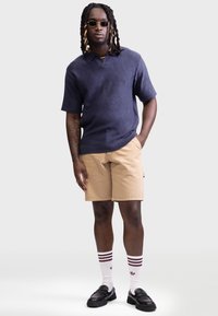 Polo navy cu mânecă scurtă, din tricot, țesătură texturată, pantaloni scurți cargo bej, mocasini negri, șosete albe cu dungi vișinii și ochelari de soare rotunzi.