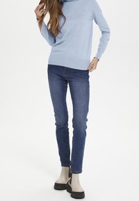 Hellblaues Strickpullover mit hohem Kragen, kombiniert mit dunkelblauen Slim-Jeans. Beigefarbene Knöchelstiefel mit einem schwarzen elastischen seitlichen Einsatz.