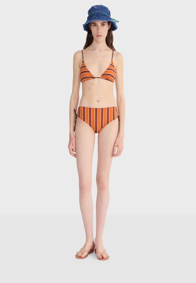 Femme portant un bikini rayé orange et marron avec des bas à nouer sur les côtés, un chapeau seau bleu et des tongs marron, debout devant un fond uni.