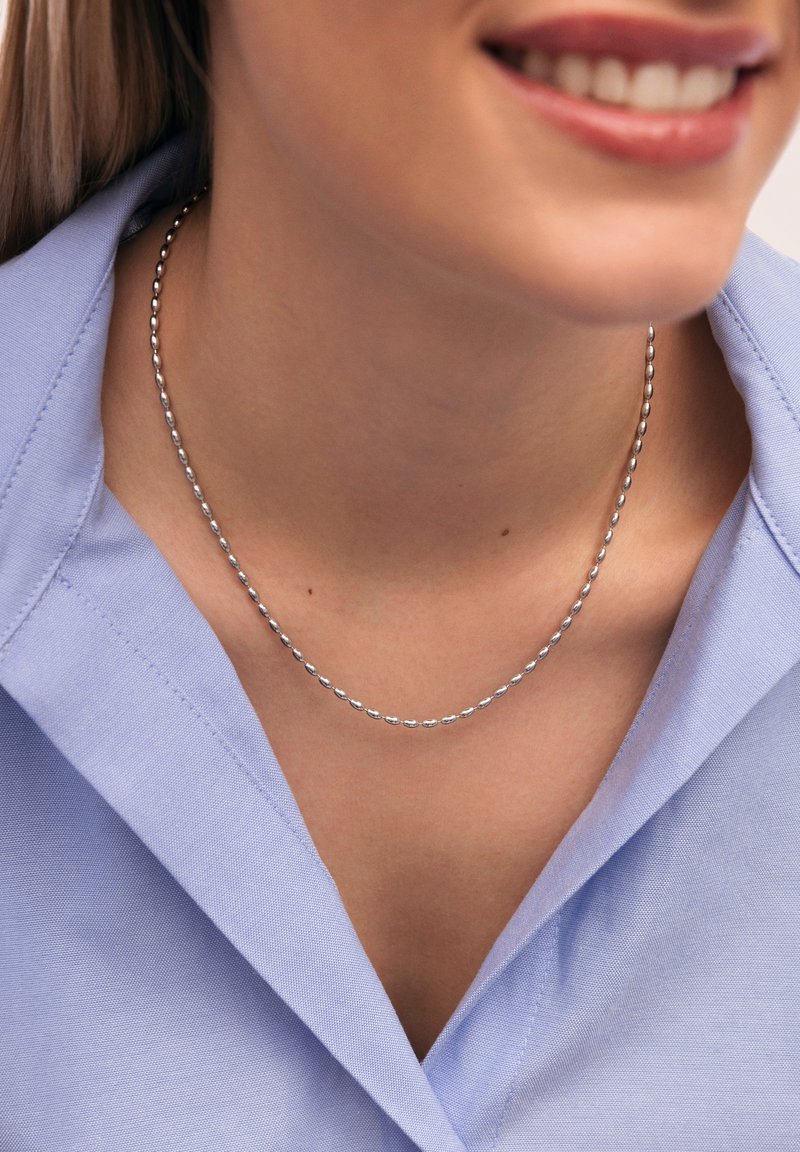 Zilveren kralenketting rustend op het sleutelbeen, gecombineerd met een lichtblauw colbert, met een soepele textuur en eenvoudig ontwerp.