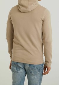Felpa beige realizzata in tessuto morbido, con cappuccio regolabile e maniche lunghe. Indossata con jeans chiari strappati.