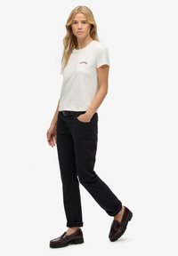 Camiseta gris de algodón con mangas cortas y detalles bordados en rojo, combinada con pantalones negros y zapatos tipo slip-on burdeos. Diseño simple.