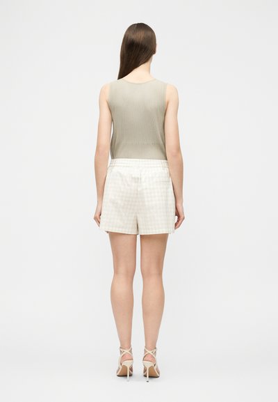 Γυναίκα που στέκεται γυρίζοντας την πλάτη, φορώντας αμάνικο μπεζ ριμπ τοπ, λευκά σορτς με καρό σχέδιο και μπεζ σανδάλια με ψηλό τακούνι.