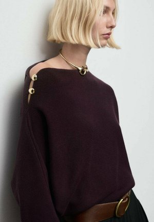 Suéter morado de gran tamaño con textura acanalada, que presenta acentos de botones dorados en un hombro, combinado con un cinturón marrón y un collar dorado.