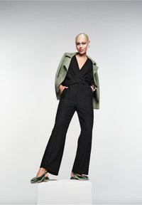 Schwarzer Jumpsuit mit Wickeldesign, grüne Wildlederjacke und grüne Mules mit Blumenverzierung. Steht auf einem Podest vor einem neutralen Hintergrund.