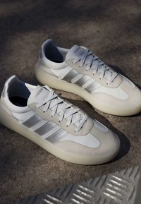 Baskets Adidas blanches et beiges avec des bandes argentées et des lacets, reposant sur une surface extérieure texturée sous une lumière douce du soleil.