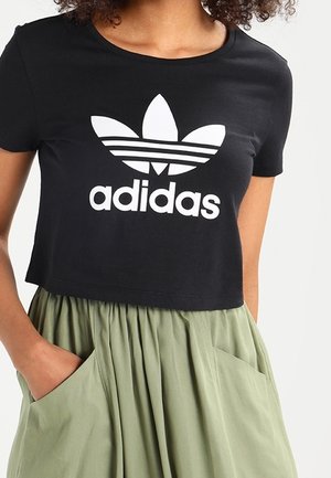 Kobieta w czarnym krótkim t-shircie Adidas i jasnozielonej spódnicy, z rękami w kieszeniach na jednolitym tle.