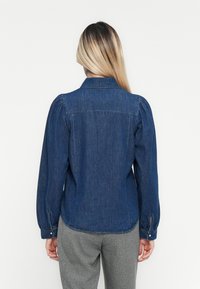 Camisa de denim em azul escuro com mangas largas bufantes, colarinho pontiagudo e punhos com botões. Apresenta uma bainha curvada e detalhes de costura subtis.