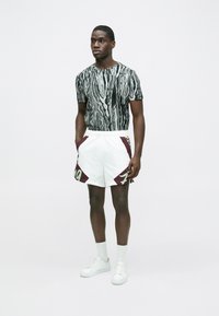 Melna un baltā rakstainā īsroku krekls kopā ar baltām šortām ar tumši sarkanām detaļām un zaļiem burtiem. Izskatu papildina baltas čības.