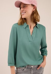 Frau in einem türkisfarbenen V-Ausschnitt-Bluse und grauen Jeans, mit einer Hand in der Tasche, lächelt und schaut zur Seite, trägt eine rosa Cordmütze.