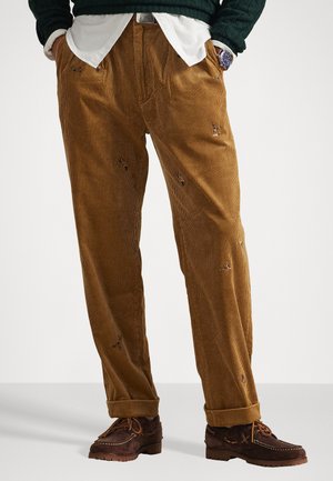 Pantalones de pana marrón con una textura acanalada, que presentan detalles bordados y parches desgastados. Combinados con zapatos de ante marrón.