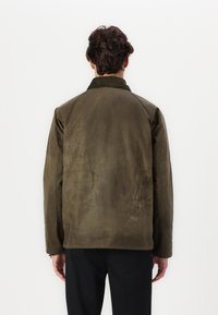 Mann, der sich abkehrt, trägt eine olivgrüne Wildlederjacke mit einem dunklen Kragen und schwarze Hosen vor einem schlichten weißen Hintergrund.