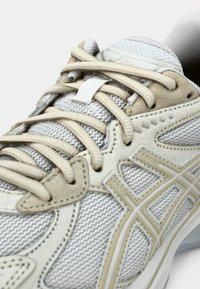Gros plan d'une chaussure de course en mesh blanc et beige avec des lacets beiges noués sur la languette rembourrée.