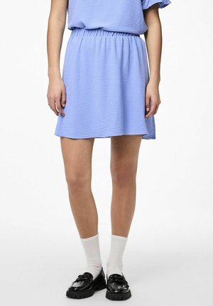 Vrouw draagt een lichtblauwe, getextureerde rok, witte geribbelde sokken en zwarte chunky loafers, staand tegen een witte achtergrond.