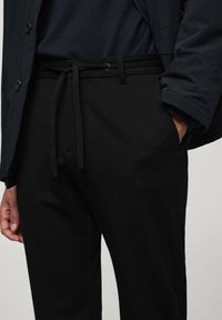 Man in zwarte drawstring broek en een donkere jas met één hand in zijn zak voor een effen achtergrond.
