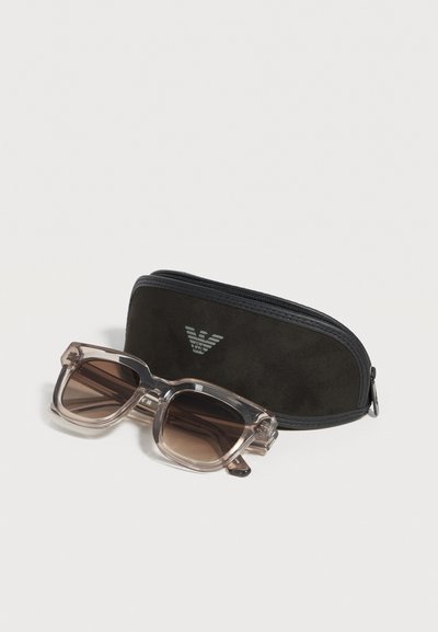 Διαφανή γκρι γυαλιά ηλίου με τετράγωνο σκελετό και καφέ φακούς, τοποθετημένα δίπλα σε μαύρη θήκη με φερμουάρ και ασημί λογότυπο.