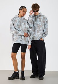 Bavlnené mikiny svetlomodrej farby s hnedým mramorovým vzorom. Model má na sebe čierne cyklistické šortky a čierne šnurovacie čižmy s hrubou podrážkou.