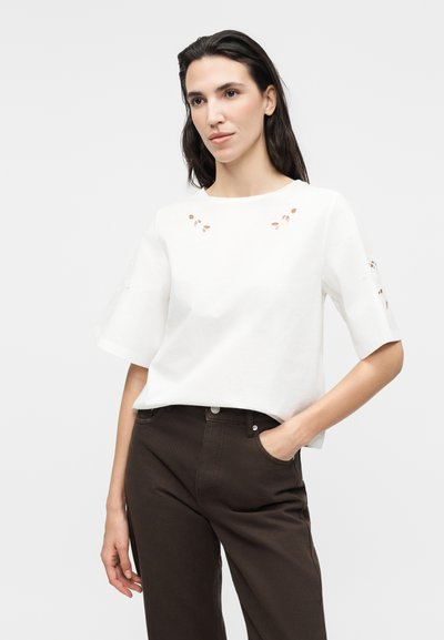 Γυναίκα με σκούρα μαλλιά που φορά λευκό μπλουζάκι με κοντά μανίκια και μικρές λεπτομέρειες κοψίματος, και σκούρο καφέ παντελόνι, χέρι στην τσέπη.