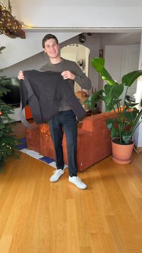 Maglione nero con trama a coste tenuto da una persona che indossa una maglia verde e pantaloni scuri, all'interno con una pianta e un divano sullo sfondo.