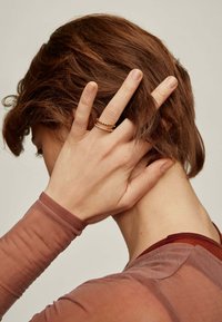 Anillos apilados en tono dorado con una banda lisa y un diseño retorcido, llevados en una mano con una blusa de manga larga transparente de color marrón claro.