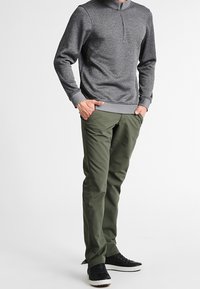 Hombre con sudadera gris de cremallera cuartilla, pantalones verdes oliva y zapatillas negras, de pie con las manos en los bolsillos contra un fondo blanco.