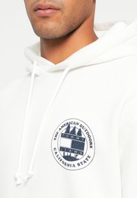 Balta džemperis su keliais mėlynais ir baltais apskritais logotipais, ant kurių užrašyta „AMERIKOS LAUKAS“ ir „KALIFORNIJOS VALSTIJA“, su raištelių uždarymu.