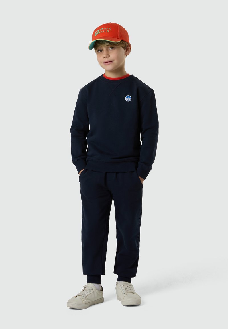 Ragazzo giovane con felpa e pantaloni blu navy, sneakers bianche e cappellino arancione North Sails, in piedi con le mani in tasca su uno sfondo semplice.