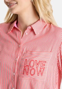 Red and white striped shirt with a collared neckline, featuring a front pocket that has "LOVE NOW" embroidered in red.