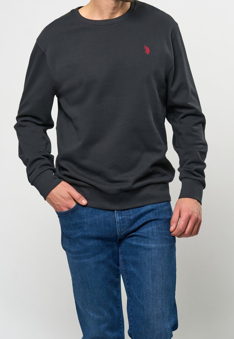 Homme portant un sweat-shirt noir uni à manches longues avec un petit logo rouge sur la poitrine et un jean bleu, main dans la poche.