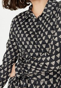 Černá košile s knoflíky, bílým vzorem připomínajícím listy a květiny, dlouhými rukávy a hedvábnou texturou. Má klasický límec a detaily s knoflíky.