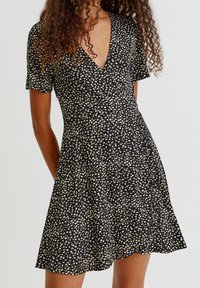 Robe noire à motifs floraux avec des manches courtes, décolleté en V, corsage ajusté, jupe évasée et motif de petites fleurs blanches et beiges partout.