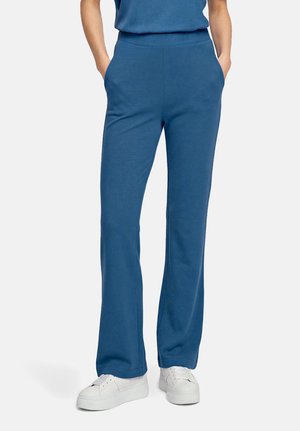 Personne portant un pantalon bleu taille haute à jambes droites, les mains dans les poches, et des baskets blanches à plateforme sur un fond uni.
