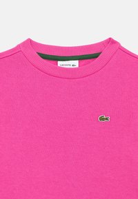 Fukszia rózsaszín pulóver bordázott, kerek nyakkivágással, bal oldalon egy kis zöld Lacoste krokodil logóval. Puha tapintású anyag.