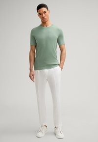 Camicetta a maglia verde chiaro a maniche corte, abbinata a pantaloni di lino bianchi semplici e sneakers bianche, in piedi contro uno sfondo neutro.
