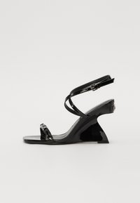 Sandale compensée en cuir verni noir avec un design élégant, à bout ouvert, des fines lanières à la cheville et une boucle décorative.